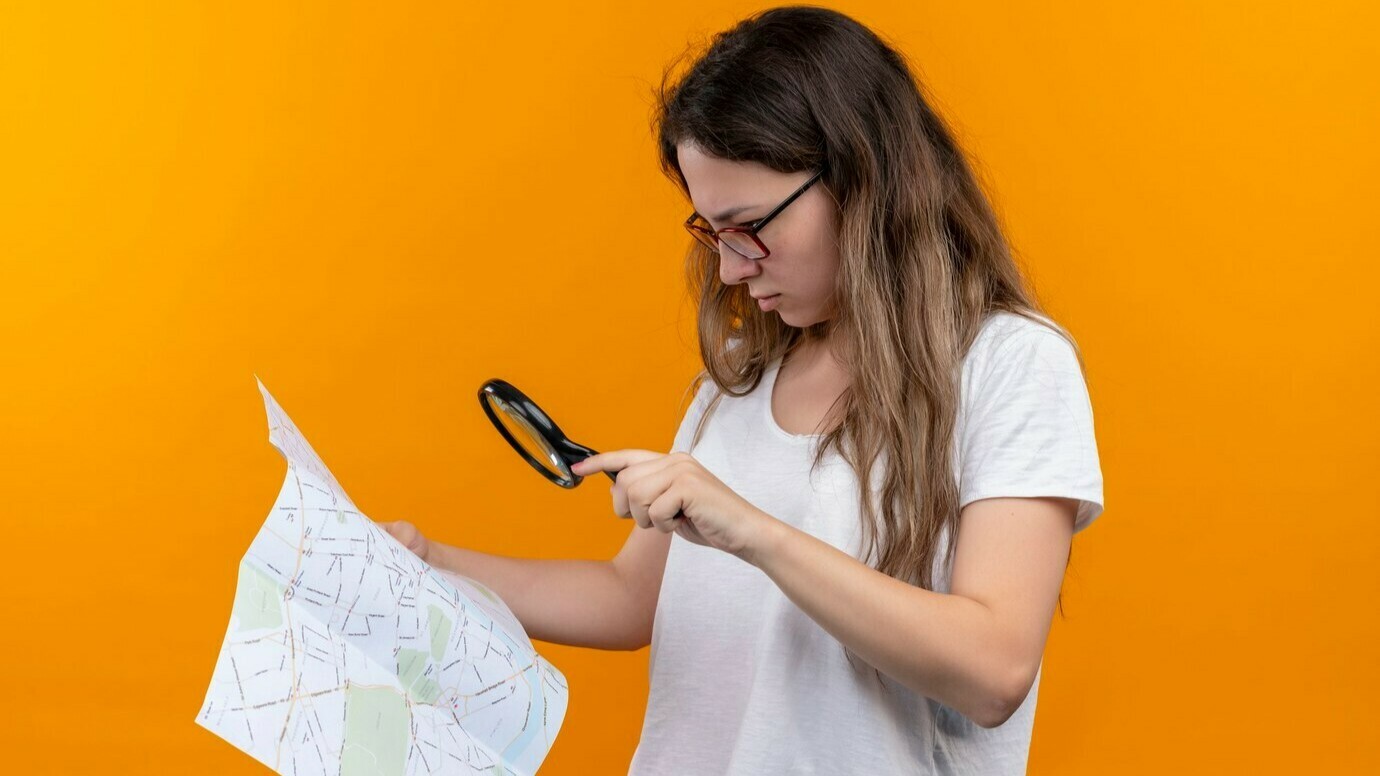 mulher-jovem-viajante-com-camiseta-branca-segurando-um-mapa-olhando-atraves-da-lupa-com-expressao-pensativa-no-rosto-pensando-em-pe-sobre-a-parede-laranja_141793-28960