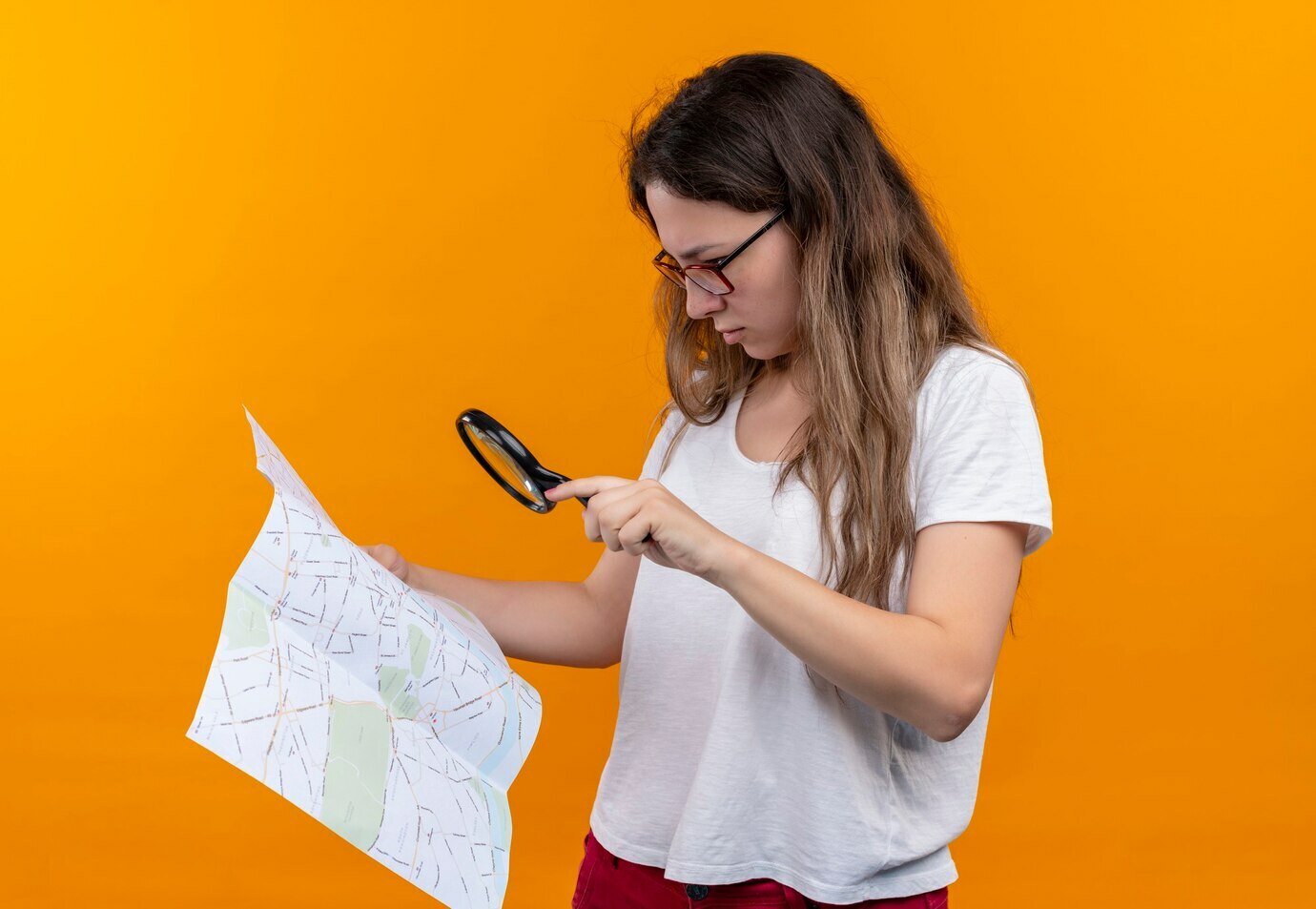mulher-jovem-viajante-com-camiseta-branca-segurando-um-mapa-olhando-atraves-da-lupa-com-expressao-pensativa-no-rosto-pensando-em-pe-sobre-a-parede-laranja_141793-28960